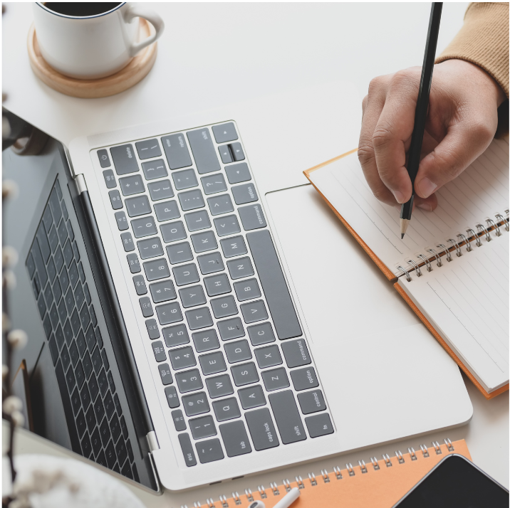 laptop and notebook on desk with a hand holding a pencil to take notes